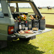 Open car trunk with picnic setup including food, wine, and flowers on a grassy field.