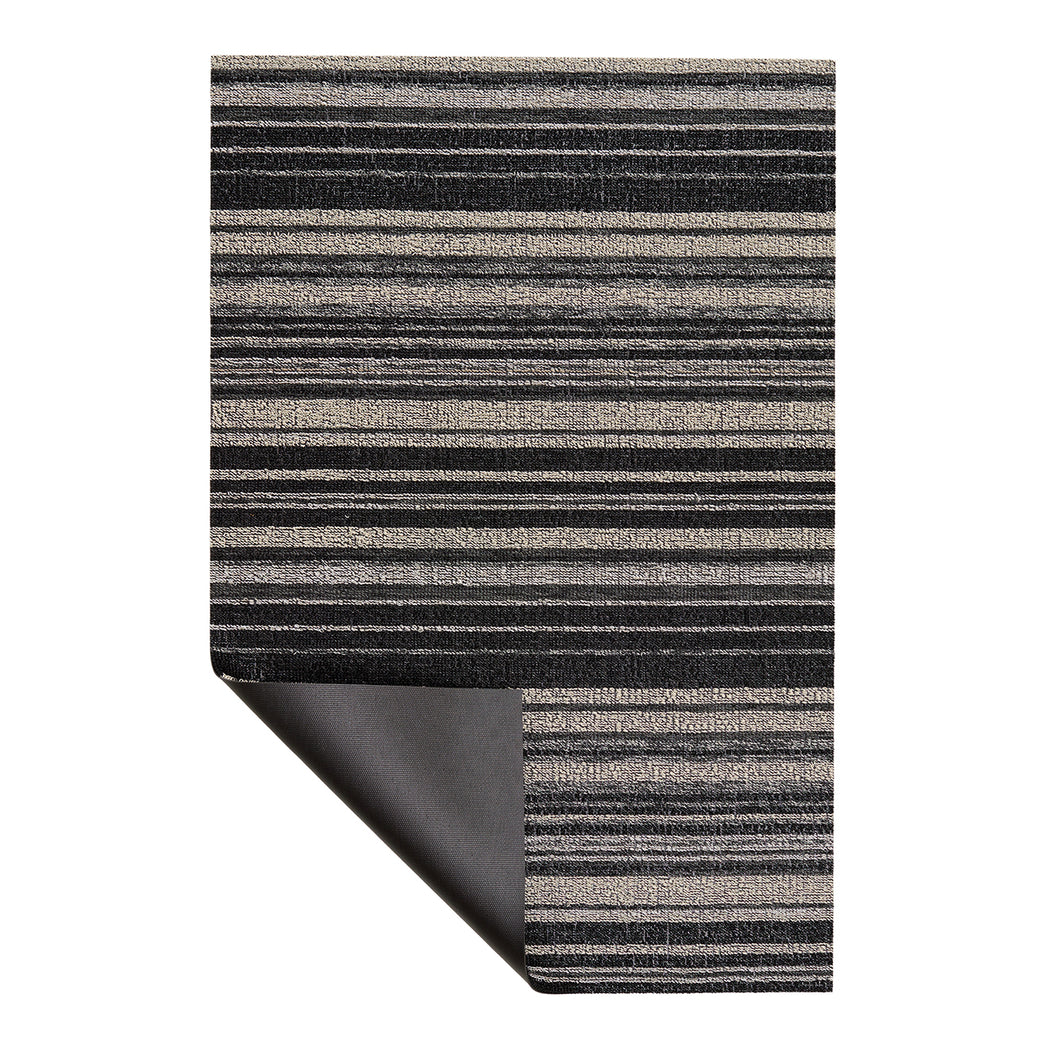 A rectangular rug with a horizontal striped pattern in black, gray, and beige, shown with one bottom corner folded over to reveal the dark backing, on a plain white background.